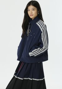 Marineblauwe zip-up jas met witte strepen en kanten accenten op de mouwen. Heeft een losse fit en een zak. Gecombineerd met een geplooide zwarte rok.