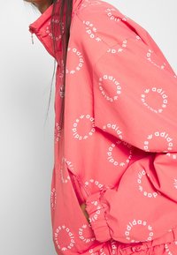 Person klädd i en korallrosa Adidas-jacka med vita cirkulära logotypmönster, sidfickor och hög krage, mot en enfärgad ljus bakgrund.