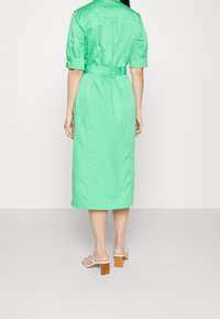 Grünes Midi-Kleid mit kurzen Ärmeln, tailliertem Bund und Rückenschlitz. Glatte Textur und minimales Design. Getragen mit beigen Blockabsatzsandalen.