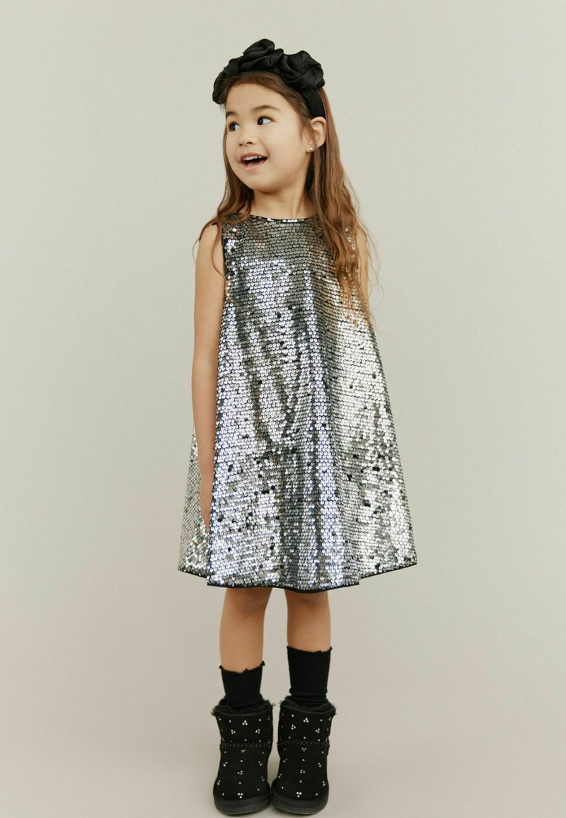 Strieborné flitrové šaty bez rukávov s voľným strihom, doplnené čiernymi ponožkami po kolená a čiernymi chlpatými topánkami zdobenými bielymi hviezdami.