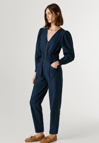 Σκούρο μπλε ολόσωμη φόρμα με βαθύ V λαιμόκοψη, φουσκωτά μανίκια και ελαστική μέση. Διαθέτει φερμουάρ μπροστά και δύο πλαϊνές τσέπες. Υφασμα απαλό.