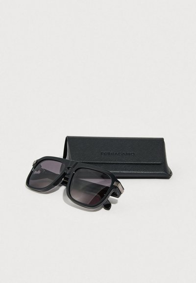 Μαύρα γυαλιά ηλίου με τετράγωνο σχήμα, φακούς με διαβάθμιση και ασημένιες λεπτομέρειες, τοποθετημένα δίπλα σε μαύρη δερμάτινη θήκη με ανάγλυφο το όνομα «Ferragamo».