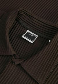 Camisa de color marrón oscuro con nervios, que presenta un cuello con solapa, un botón lateral y una etiqueta con la marca "JACK & JONES CORE" cerca del escote.