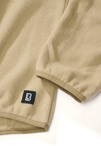 Hellbrauner Fleece-Stoff mit strukturierter Oberfläche, der am unteren Saum ein schwarzes Etikett mit weißem Logo trägt. Der Ärmel zeigt ein gerafftes Design.