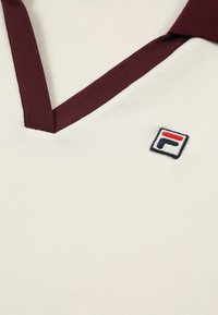 Witte polo met een bordeauxrode kraag, voorzien van een klein geborduurd logo in het blauw, rood en wit. Katoenen materiaal met een gladde textuur.