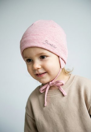 Toddler with light hair wearing a beige sweater and a pink hat tied under the chin, smiling softly against a light background.