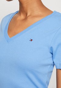 Světle modré bavlněné tričko s výstřihem do V a krátkými rukávy, s malým logem v červené, bílé a námořnické modři poblíž hrudi. Hladký materiál.