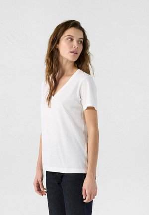Camiseta de algodón blanca con escote en V y mangas cortas, que presenta una textura suave y un corte relajado. Llevada con jeans oscuros.
