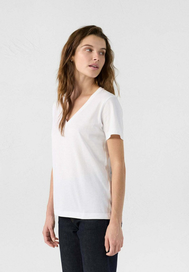 T-shirt bianco in cotone con scollatura a V e maniche corte, caratterizzato da una texture morbida e una vestibilità comoda. Indossato con jeans scuri.