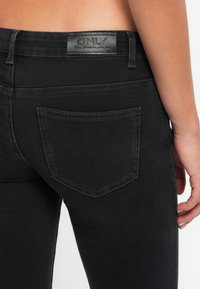 Acercamiento de una persona que lleva jeans negros con un bolsillo trasero visible y una etiqueta de cuero de la marca que dice "ONLY" en la cinturilla.