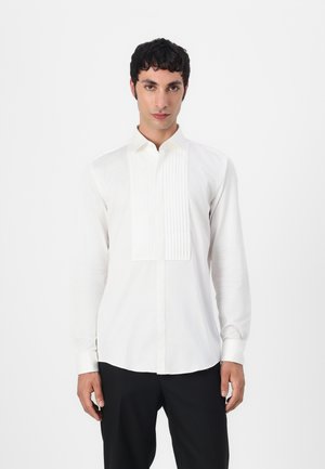 Camisa blanca de manga larga con detalle de frente con pliegues, cuello abotonado y ajuste slim. Tejido suave con textura lisa. Combinada con pantalones negros.