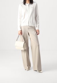 Bílý pletený cardigan s kulatým výstřihem a knoflíky, doplněný béžovými kalhotami s širokými nohavicemi. Drží světlou kabelku s řetízkem.