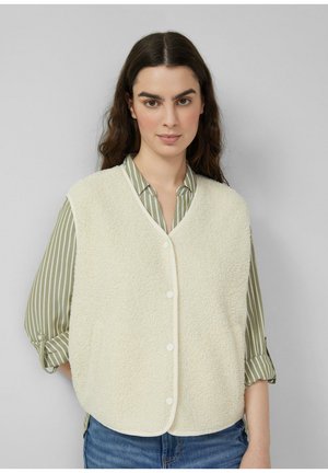 Kobieta ubrana w kremową kamizelkę polarową na guziki, na zielono-białej paskowanej koszuli i niebieskich dżinsach, stojąca na tle szarego tła.