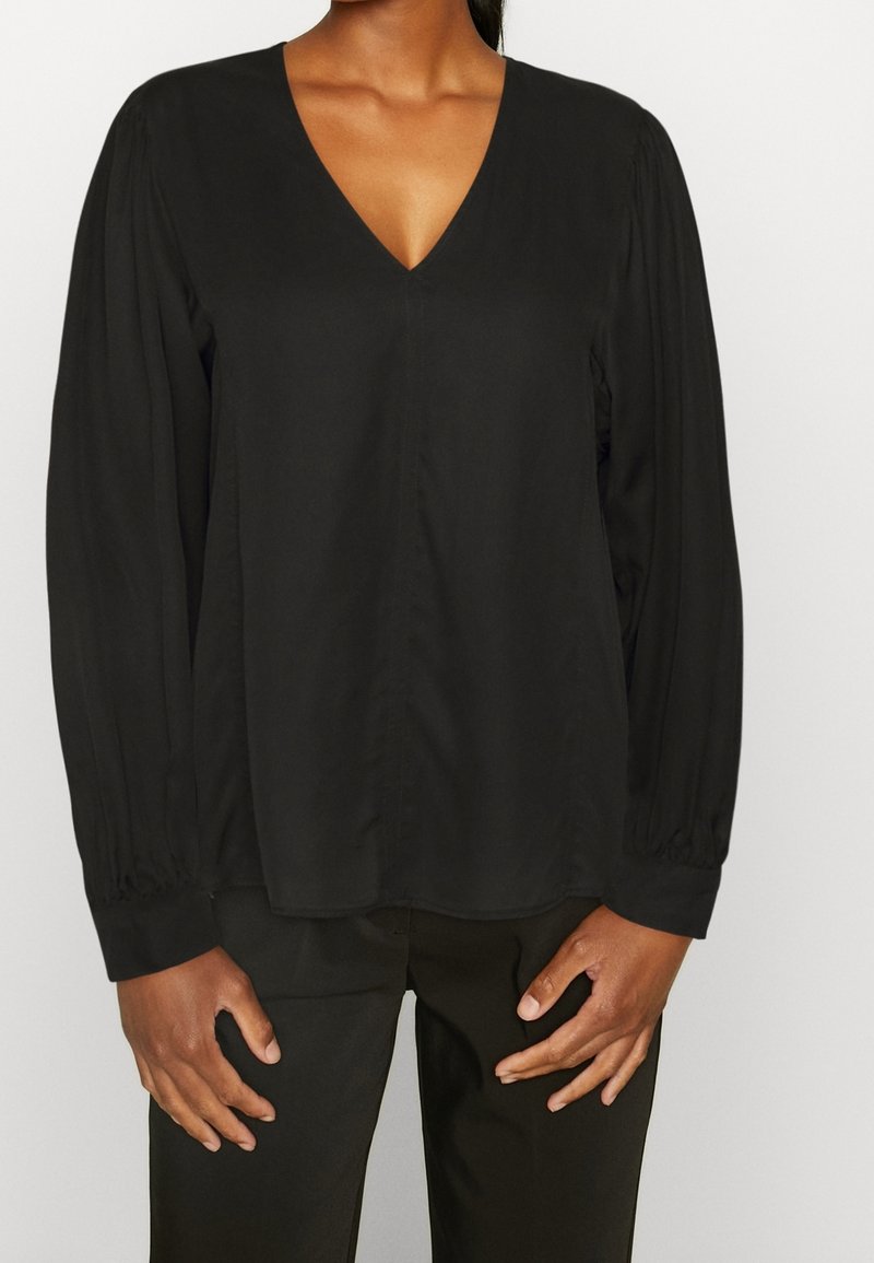 Femme portant un chemisier noir uni à manches longues avec un col en V et un pantalon noir, debout devant un fond clair uni.