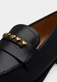 Μαύρο δερμάτινο μοκασίνι με χρυσά καρδιακά καρφάκια στο πάνω μέρος, ένα από τα οποία είναι χαραγμένο με "LOVE MOSCHINO", και λεπτομερής ραφή.