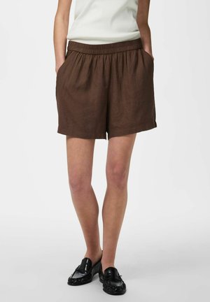 Frau trägt braune lockere Shorts mit elastischem Bund, weißes ärmelloses Oberteil und schwarze Loafers, steht mit den Händen in den Taschen.