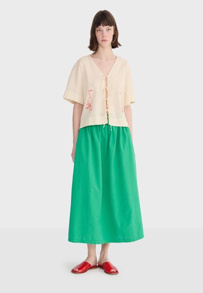 Młoda kobieta ubrana w kremową, krótkorękawową bluzkę ze sznureczkowymi zapięciami i czerwonym haftem przedstawiającym ptaki, zieloną spódnicę midi oraz czerwone sandały z odkrytymi palcami.