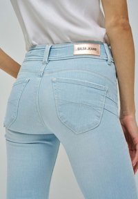 Vaqueros de mezclilla azul claro con corte ajustado, dos bolsillos traseros y una etiqueta de marca metálica "SALSA JEANS" en la cinturilla.