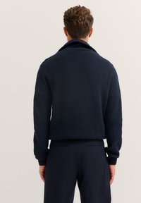 Dunkelblaue Strickjacke mit gerippter Textur, umgeschlagenem Kragen und abfallenden Schultern, kombiniert mit dunklen Hosen und einer figurschmeichelnden Silhouette.