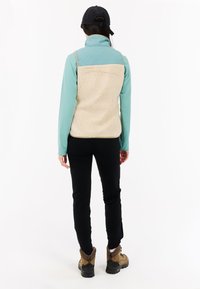 Lichtgewicht fleece vest met een gevoerde turquoise bovenkant en een crèmekleurige textuur onderaan. Draagt over zwarte broeken en bruine wandelschoenen.