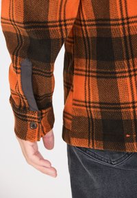 Orange og sort plaid flannel skjorte med knapmanchetter, brunt tekstureret stof og grå accenter nær ærmerne. Synlig håndbevægelse.