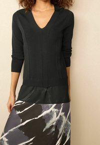 Schwarzer, ribbeliger Pullover mit V-Ausschnitt und langen Ärmeln, über einem dunklen, transparenten Top und einem gemusterten, fließenden Rock in Grautönen und Schwarz getragen.