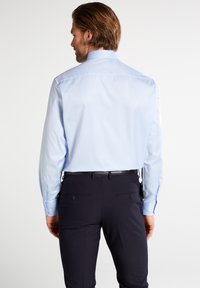 Camicia a maniche lunghe di colore azzurro chiaro realizzata in tessuto morbido, con colletto standard e polsini con bottone, indossata con pantaloni scuri.