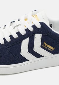 Námořnické semišové tenisky s bílou gumovou podrážkou, se stříbrnými akcenty a zlatým logem. Bělé tkaničky dokončují design.
