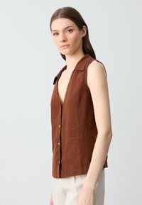 Camicia senza maniche in lino marrone con scollatura a V, chiusura frontale a bottoni e una vestibilità leggermente ampia. Texture morbida e design minimalista.