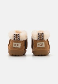 Brune suède ankelstøvler med fluffy kremfarge pelskrage, teksturert gummisåle og vevde brune og hvite aksenter på baksiden. UGG-logoen er synlig.