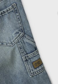 Tela de mezclilla de color azul claro con un diseño de bolsillo en diagonal. Presenta una etiqueta de marca marrón con detalles de costura y textura visible.