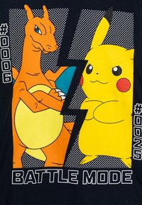 Graphiques d'un Charizard orange et d'un Pikachu jaune sur un t-shirt navy, avec un texte en gras "MODE BATAILLE" et un fond à pois.