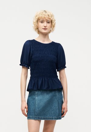 Femme blonde aux cheveux bouclés portant un chemisier bleu marine à manches courtes froncées et une mini-jupe en denim bleu, debout devant un fond blanc.