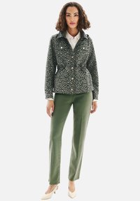 Veste à imprimé léopard vert olive avec fermeture à boutons, taille réglable, associée à un pantalon vert à coupe droite et des chaussures pointues beige.