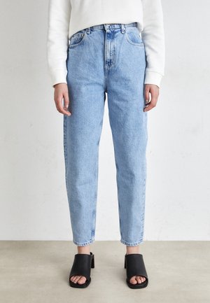 Personne portant un jean taille haute bleu clair, un haut blanc à manches longues et des chaussures noires à talons ouverts, debout sur un sol clair.