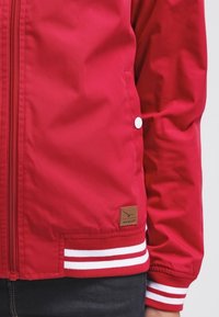 Veste rouge avec poche à bouton-pression blanche, fermeture éclair frontale, ourlet côtelé et poignets avec des rayures blanches, et une petite écusson logo marron.