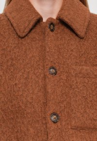 Giacca camicia in misto lana marrone con una finitura testurizzata, caratterizzata da un colletto classico, chiusura con bottoni e una tasca a soffietto sul lato sinistro.