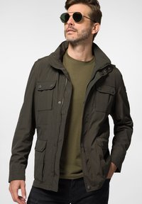 Oliwkowa kurtka z kapturem, zapinana na zamek, z wieloma kieszeniami z przodu. Wykonana z lekkiego materiału o gładkiej teksturze, zestawiona z zieloną koszulką.