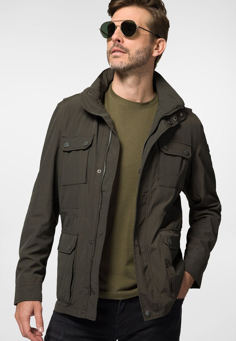Oliwkowa kurtka z kapturem, zapinana na zamek, z wieloma kieszeniami z przodu. Wykonana z lekkiego materiału o gładkiej teksturze, zestawiona z zieloną koszulką.