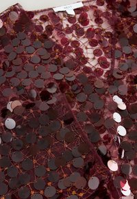 Burgundy fabric top with a sheer base, embellished with circular sequins and gold thread detailing, creating a textured design.