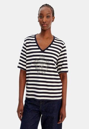 Giovane donna in piedi, che indossa una maglietta a righe orizzontali bianche e nere con scollo a V e un disegno a stemma, abbinata a jeans blu scuro su uno sfondo semplice.