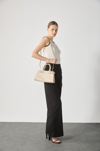 Beige håndveske med en strukturert design og doble håndtak. Modellen har på seg en hvit topp med sorte detaljer og svarte vide bukser, mot en nøytral bakgrunn.