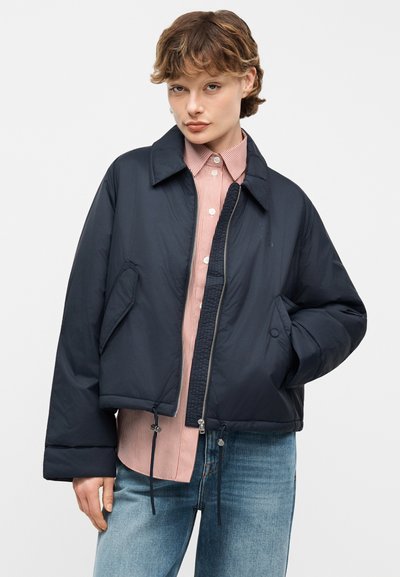 Νεαρό άτομο που φοράει μπλε σκουρόχρωμο μπουφάν τύπου puffer πάνω από πουκάμισο με κόκκινες ρίγες και μπλε τζιν, στέκεται με το ένα χέρι στην τσέπη.