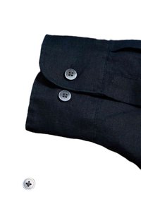 Polsino di camicia nera con due bottoni rotondi grigi; il tessuto sembra testurizzato con cuciture visibili. Un singolo bottone è posizionato accanto al polsino.