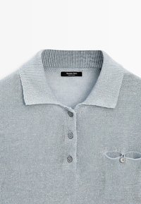 Polo in maglia di colore grigio chiaro con colletto a coste, tre bottoni sul davanti e una piccola tasca con bottone sul petto.