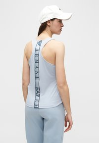 Γυναίκα που φοράει αμάνικη γαλάζια μπλούζα με κάθετο λογότυπο "Armani EA7" στην πλάτη, παντελόνια που ταιριάζουν και λευκό καπέλο μπέιζμπολ, κοιτάζοντας πλαγίως.