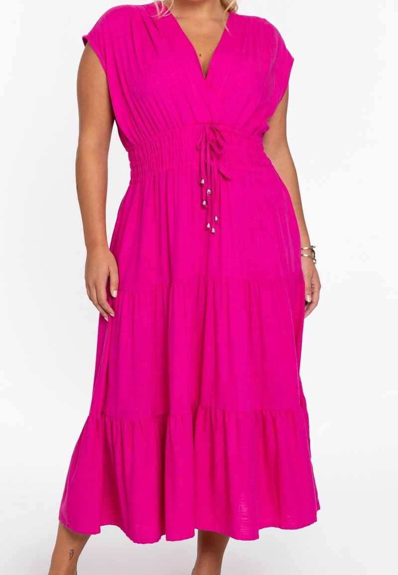 Femme portant une robe midi rose vif avec manches courtes, taille froncée avec cordon et jupe à volants sur fond blanc uni.