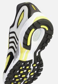 Svart, grå och vit sportskor med gula accenter. Har en texturerad ovandel, mesh-paneler och en mönstrad gummisula.