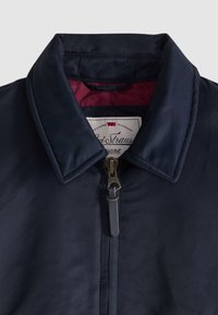 Γιακάς navy blue μπουφάν με φερμουάρ, με δερμάτινη λαβή φερμουάρ και εμφανή ετικέτα εσωτερικού με κόκκινη επένδυση.