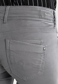 Pantalones de mezclilla grises con un bolsillo trasero que presenta costuras sutiles y un acento de botón metálico. El material es suave con un acabado ligeramente mate.